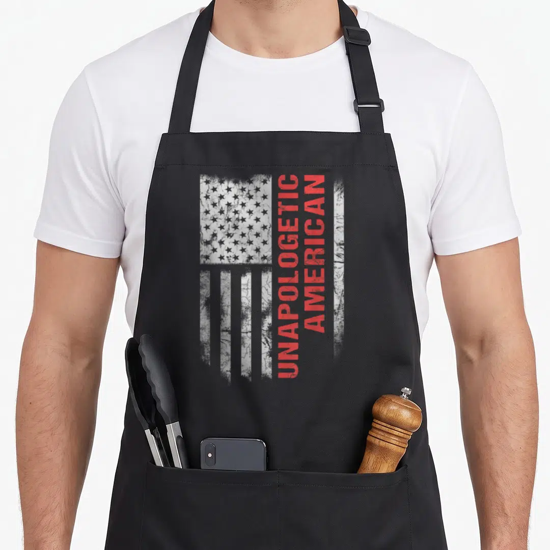 Unapologetic veterans day patriotic american flag apron - Vertical red UNAPOLOGETIC AMERICAN text over distressed grey — shown on a black cotton canvas apron against a white background