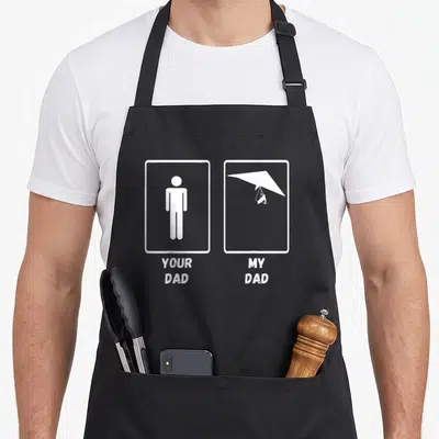 Hang glider apron - Two white frames showing a stick figure and hang glider with text YOUR DAD MY DAD — shown on a black cotton canvas apron against a white background