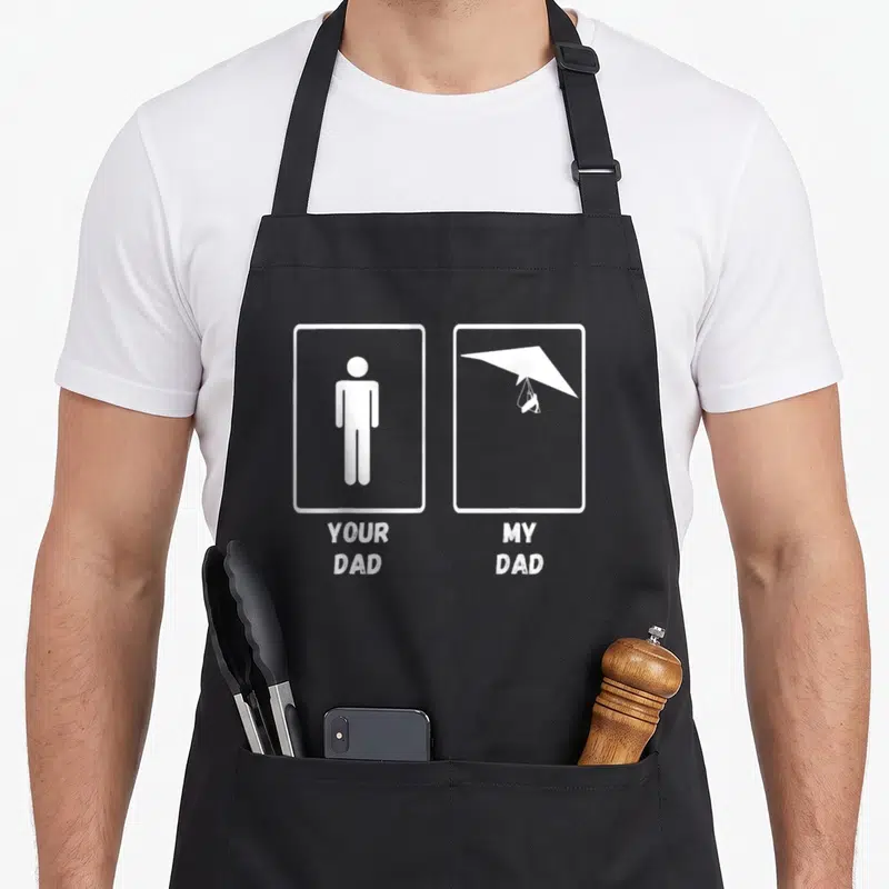 Hang glider apron - Two white frames showing a stick figure and hang glider with text YOUR DAD MY DAD — shown on a black cotton canvas apron against a white background