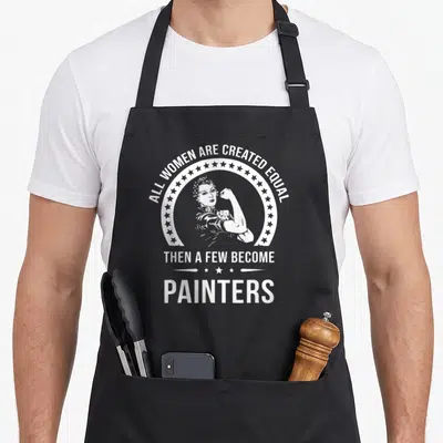 Funny painter apron - Retro woman flexing arm in star circle with white text about women becoming painters — shown on a black cotton canvas apron against a white background