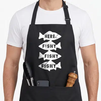 Fish fishing bait apron - Four white fish silhouettes stacked vertically containing HERE FISHY FISHY FISHY text — shown on a black cotton canvas apron against a white background