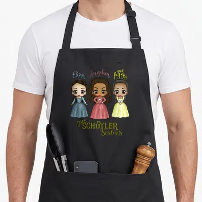 Schuyler kawaii apron - Three cartoon women in blue pink and yellow dresses with Eliza Angelica Peggy text — shown on a black cotton canvas apron against a white background