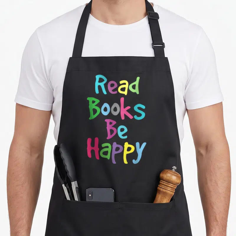 Reading literacy teacher apron - Stacked colorful text reading Read Books Be Happy in blue, yellow, green, pink — shown on a black cotton canvas apron against a white background