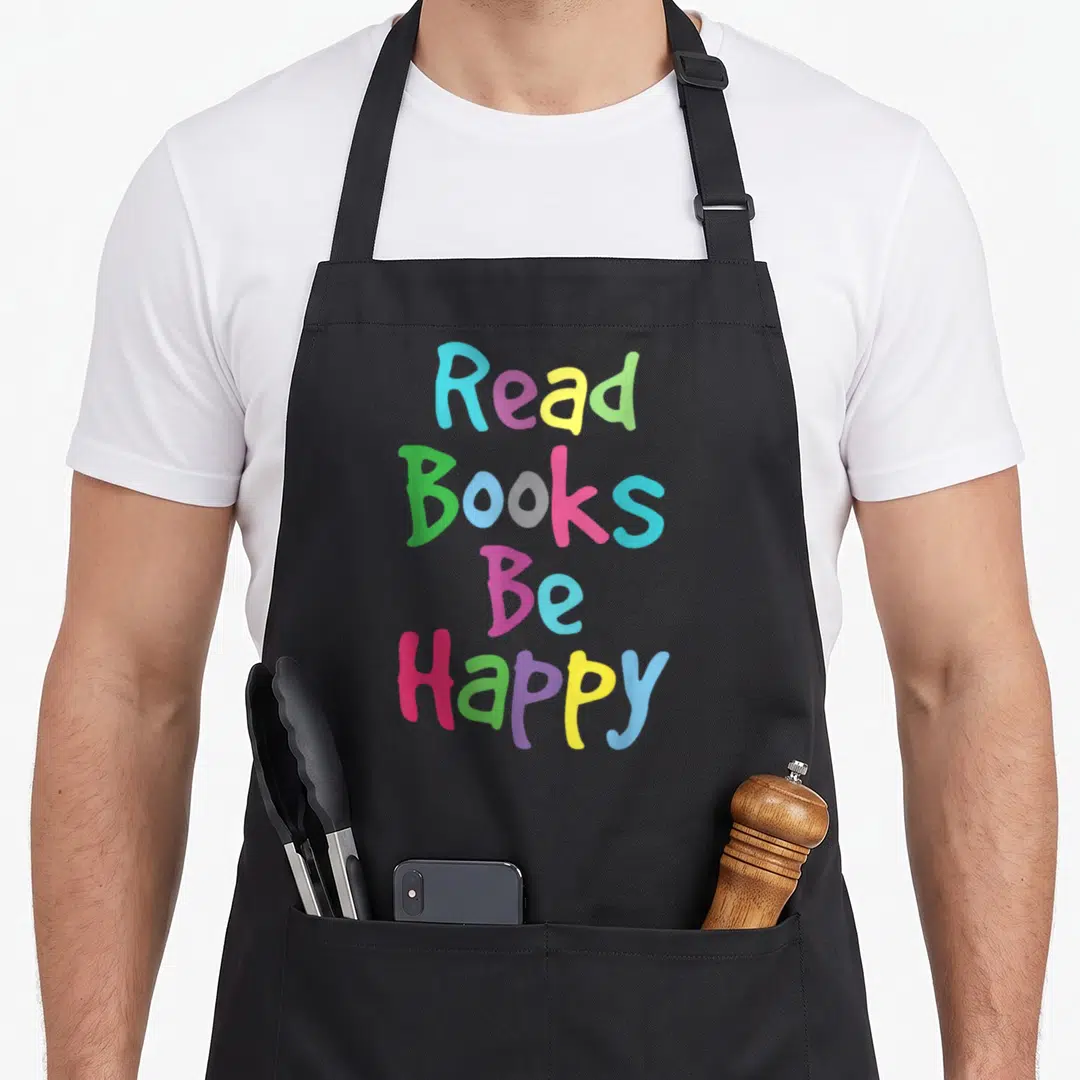 Reading literacy teacher apron - Stacked colorful text reading Read Books Be Happy in blue, yellow, green, pink — shown on a black cotton canvas apron against a white background