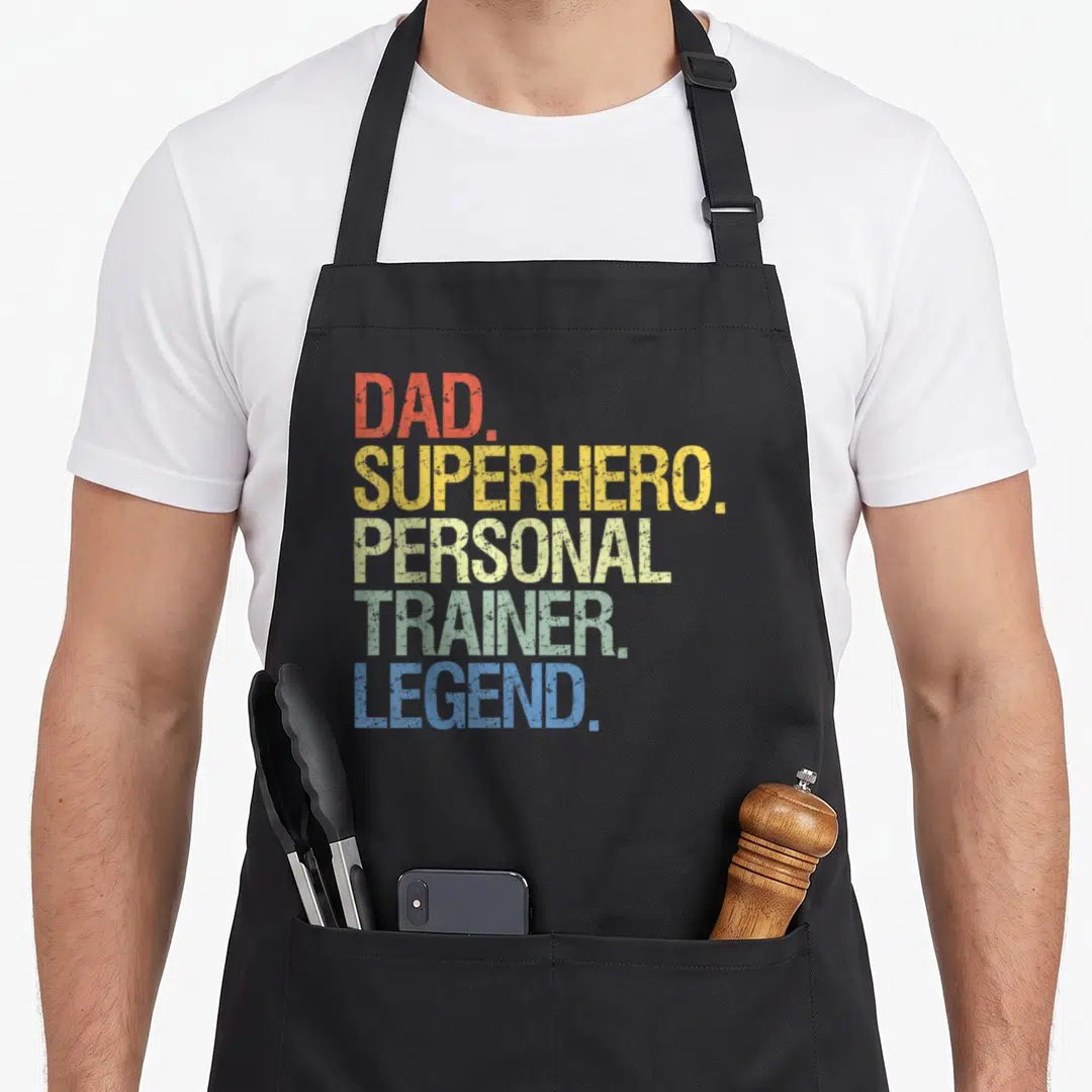 Trainer personal printed aprons - Stacked DAD SUPERHERO PERSONAL TRAINER LEGEND text in red yellow green blue — shown on a black cotton canvas apron against a white background