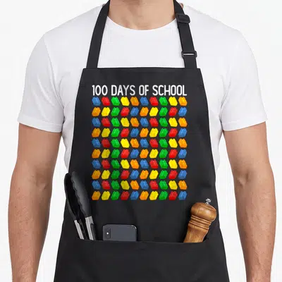 School building apron - 100 DAYS OF SCHOOL text above 100 colorful building blocks in a grid pattern — shown on a black cotton canvas apron against a white background