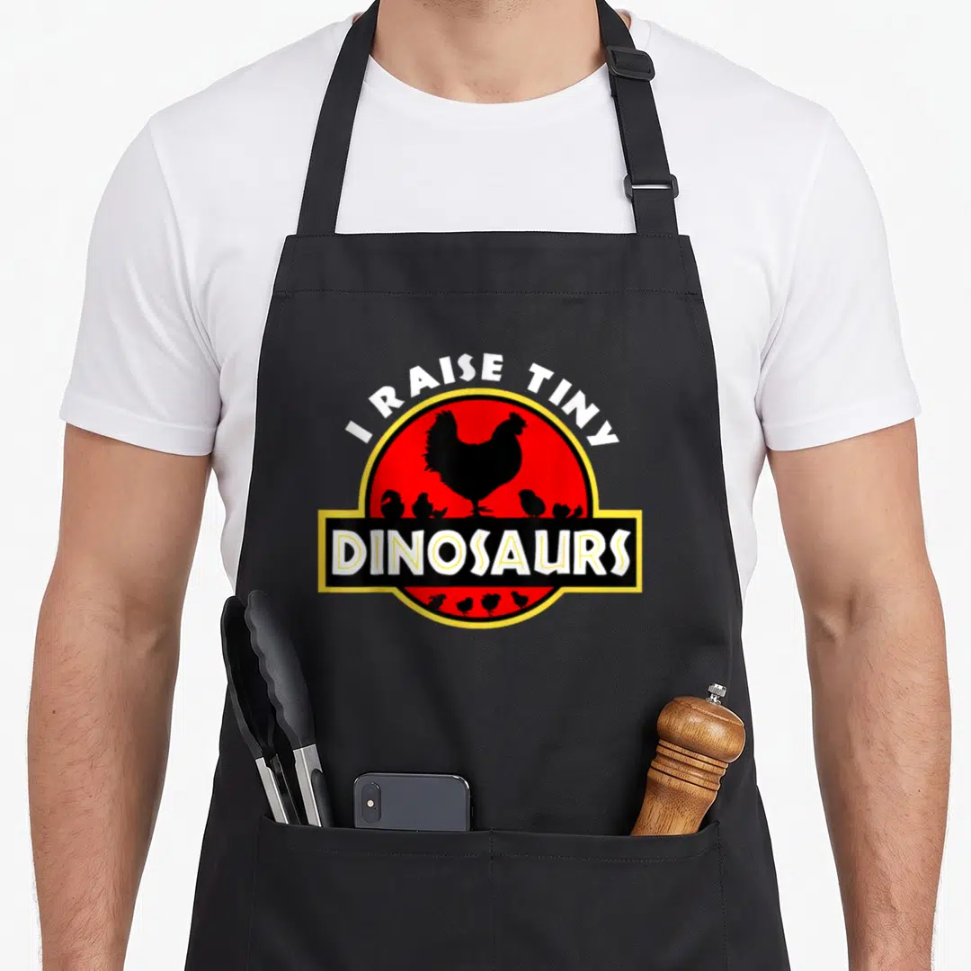 Raise tiny chef apron - I RAISE TINY DINOSAURS text over red circle with black chicken and chick silhouettes — shown on a black cotton canvas apron against a white background