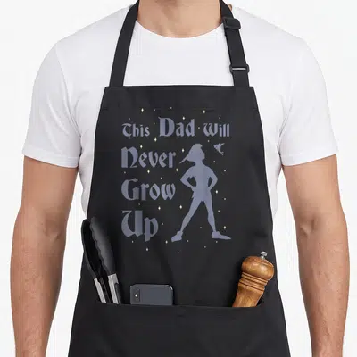 Peter robin dad apron - Purple text This Dad Will Never Grow Up beside Peter Pan silhouette and yellow stars — shown on a black cotton canvas apron against a white background