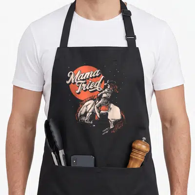 Mama western aprons - Retro illustration of a woman riding a bucking horse next to stacked Mama Tried text inside — shown on a black cotton canvas apron against a white background