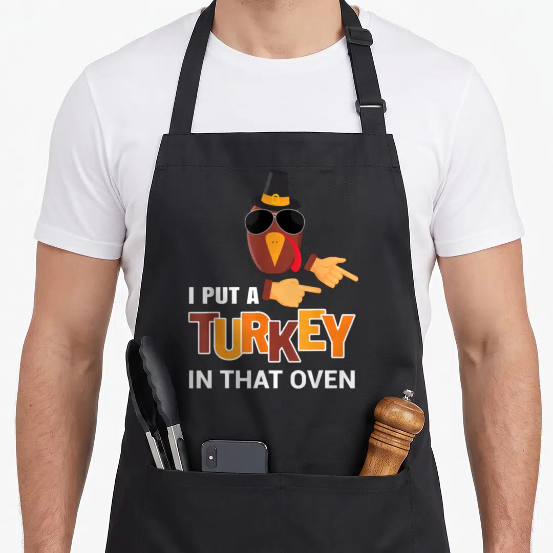 Funny pregnancy thanksgiving apron - Cartoon turkey pointing to I PUT A TURKEY IN THAT OVEN text in brown orange white — shown on a black cotton canvas apron against a white background