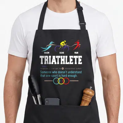 Triathlon humorous aprons - Three colored athletes swimming biking running above dictionary definition text — shown on a black cotton canvas apron against a white background