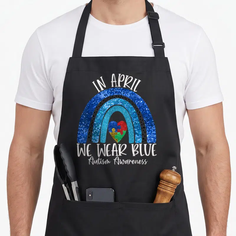Support autism acceptance apron - Blue glitter rainbow over puzzle heart with we wear blue text — shown on a black cotton canvas apron against a white background