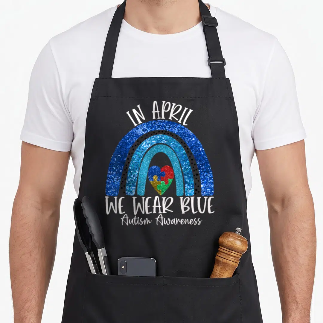 Support autism acceptance apron - Blue glitter rainbow over puzzle heart with we wear blue text — shown on a black cotton canvas apron against a white background