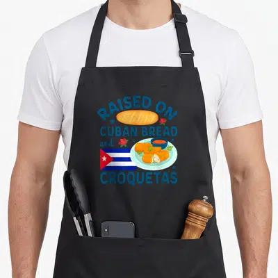 Cuban bread apron - Raised On Cuban Bread and Croquetas text with loaf, plate, and flag in blue orange red white — shown on a black cotton canvas apron against a white background
