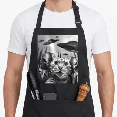Peace funny cat apron - Tabby cat selfie with two grey aliens and UFOs over trees in grayscale — shown on a black cotton canvas apron against a white background
