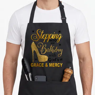 Stepping grace apron - Gold glitter high heel shoe with stacked black and gold birthday faith text and stars — shown on a black cotton canvas apron against a white background