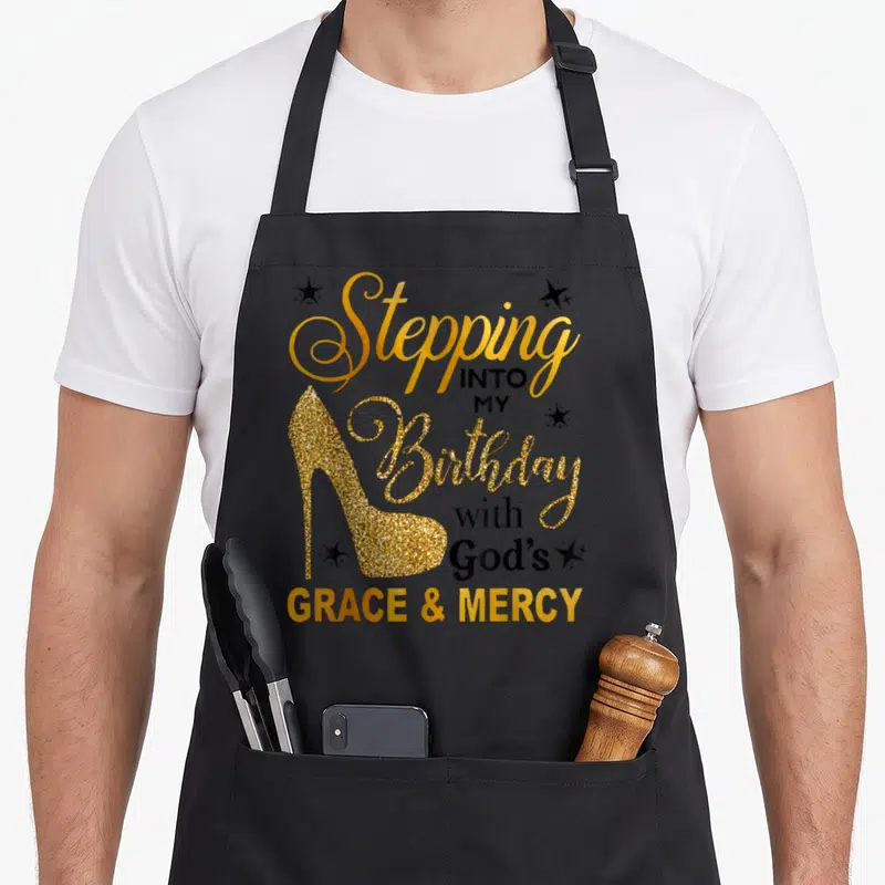 Stepping grace apron - Gold glitter high heel shoe with stacked black and gold birthday faith text and stars — shown on a black cotton canvas apron against a white background