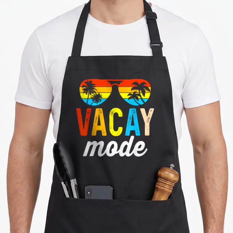 Vacation holiday apron - Retro sunglasses with palm tree sunset reflection and VACAY mode text in red yellow blue — shown on a black cotton canvas apron against a white background