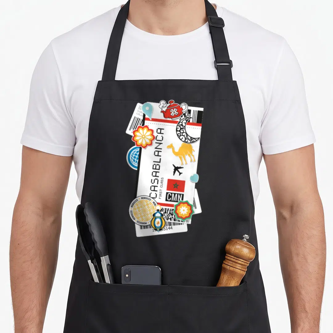 Casablanca travel aprons - White ticket with CASABLANCA text, yellow camel, red teapot, and moon icons in cartoon style — shown on a black cotton canvas apron against a white background