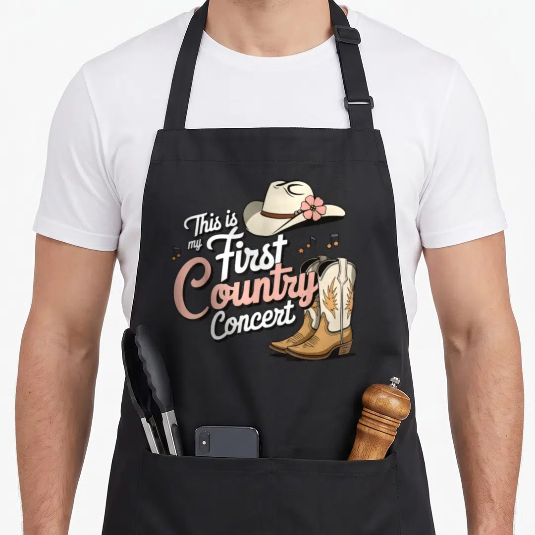 Cute concert country kitchen apron - Text 'This is my First Country Concert' with cowboy hat, boots, and musical notes — shown on a black cotton canvas apron against a white background