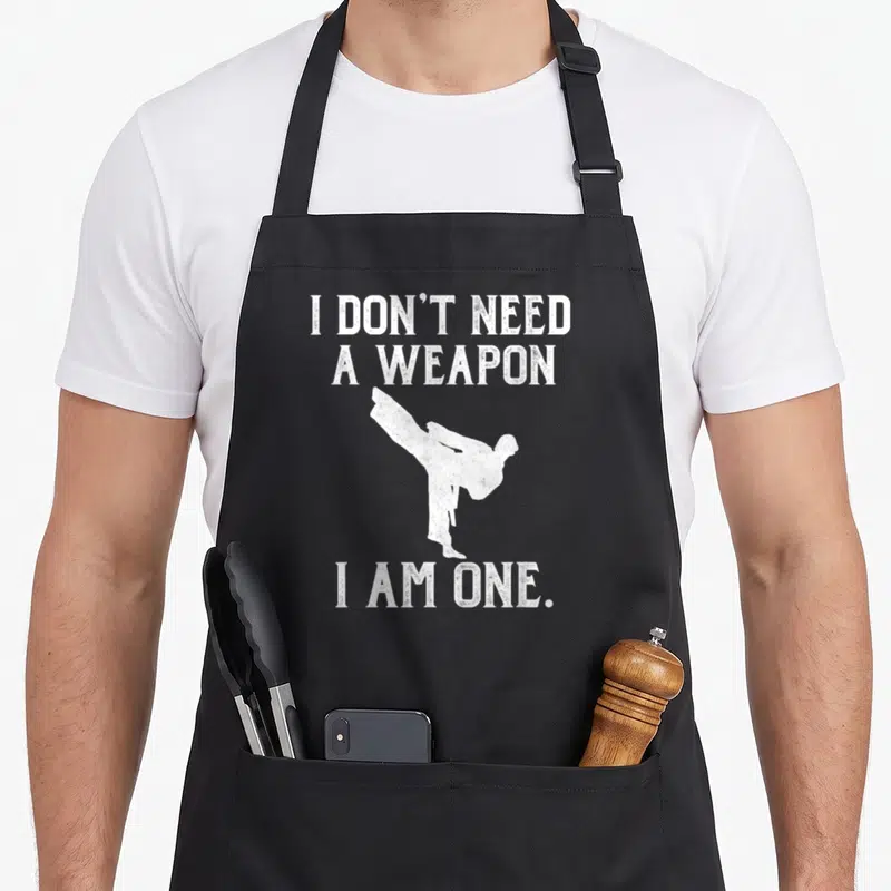 Weapon kung apron - White martial artist kicking silhouette with distressed text I don't need a weapon I am one — shown on a black cotton canvas apron against a white background