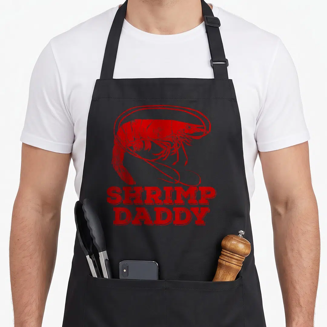 Shrimp daddy apron - Red distressed shrimp inside circle above stacked SHRIMP DADDY text — shown on a black cotton canvas apron against a white background