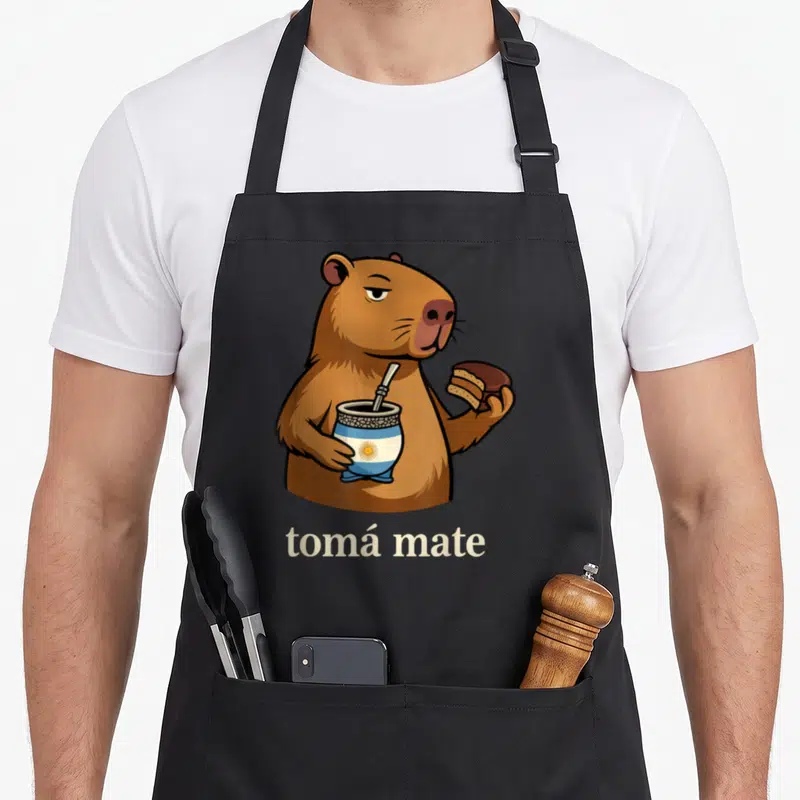 Argentinian alfajores apron - Cartoon capybara holding striped mate cup and alfajor with tomá mate text below — shown on a black cotton canvas apron against a white background