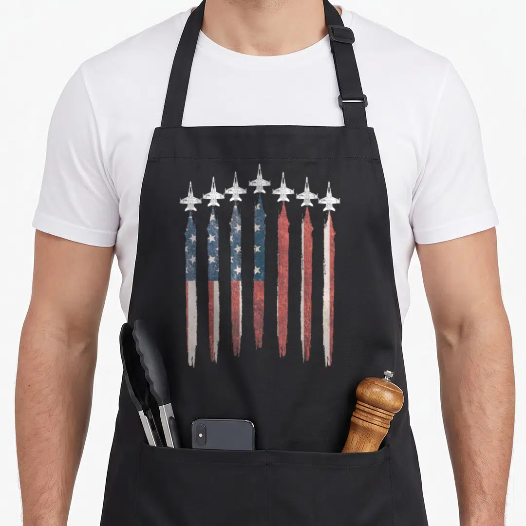 F18 4th of july apron - Seven fighter jets leaving smoke trails that form a distressed red, white, and blue flag — shown on a black cotton canvas apron against a white background