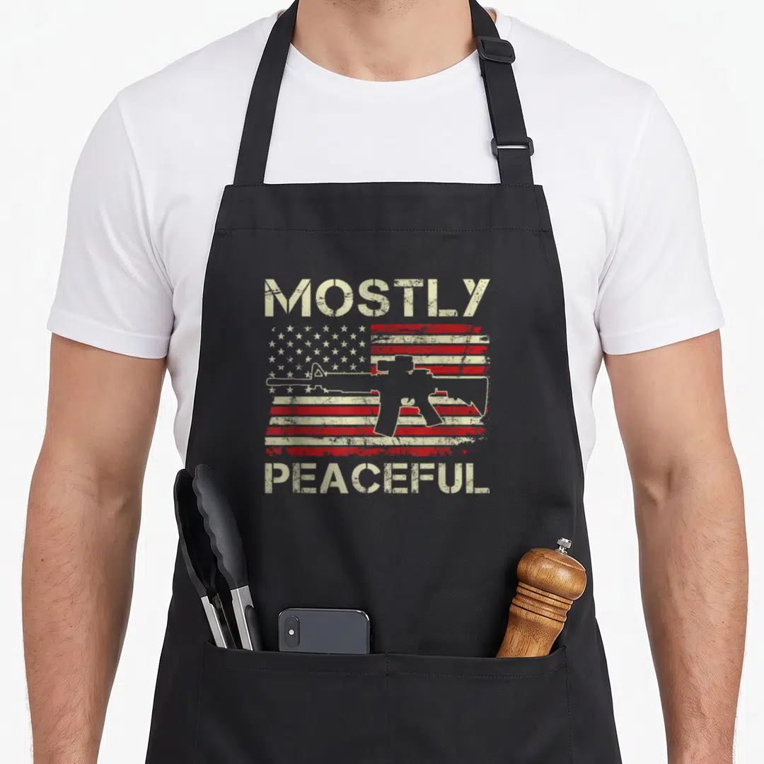 Mostly 2nd apron - Distressed USA flag with rifle silhouette and text MOSTLY PEACEFUL in beige red and white — shown on a black cotton canvas apron against a white background