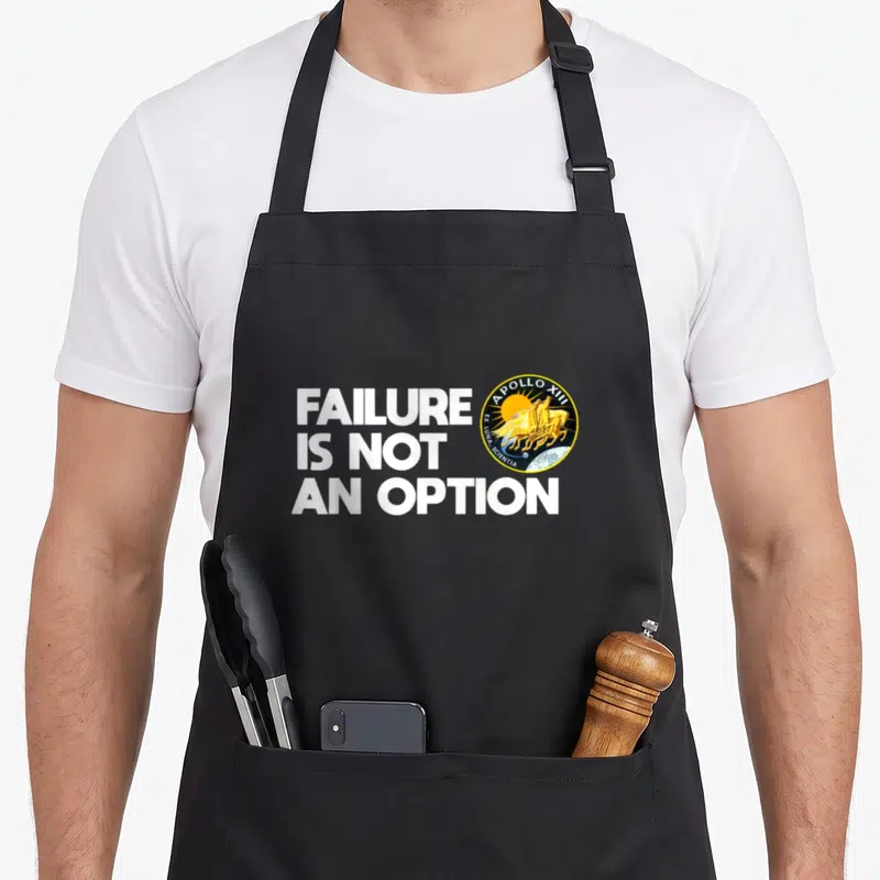 Option nasa apron - Failure Is Not An Option text next to Apollo 13 mission patch with yellow horses — shown on a black cotton canvas apron against a white background