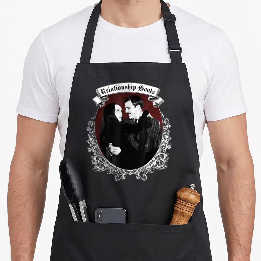 Relationship goth apron - Couple embracing in dark red oval with Relationship Goals text on white ribbon — shown on a black cotton canvas apron against a white background