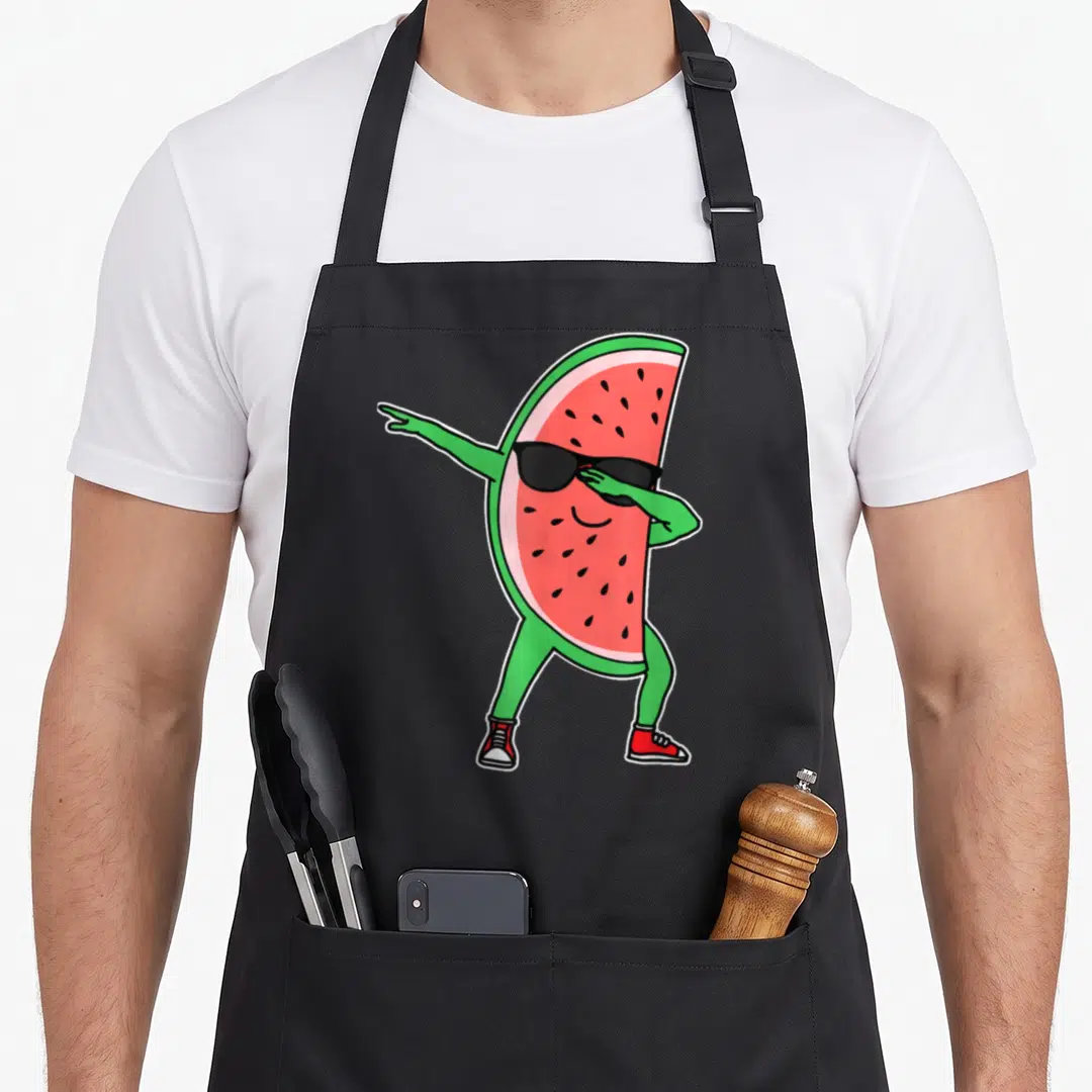 Kawaii watermelon apron - Dabbing cartoon watermelon slice with sunglasses and red sneakers in red and green colors — shown on a black cotton canvas apron against a white background