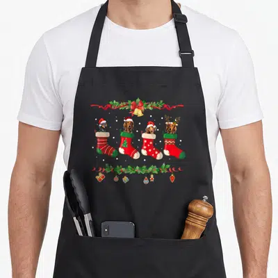 Dachshund christmas dog apron - Four cartoon dachshunds in red and green stockings framed by holly ribbons and hanging — shown on a black cotton canvas apron against a white background