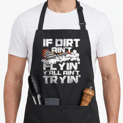 Flyin track apron - Distressed white text IF DIRT AIN'T FLYIN YALL AIN'T TRYIN over checkered flag with fire and dust — shown on a black cotton canvas apron against a white background