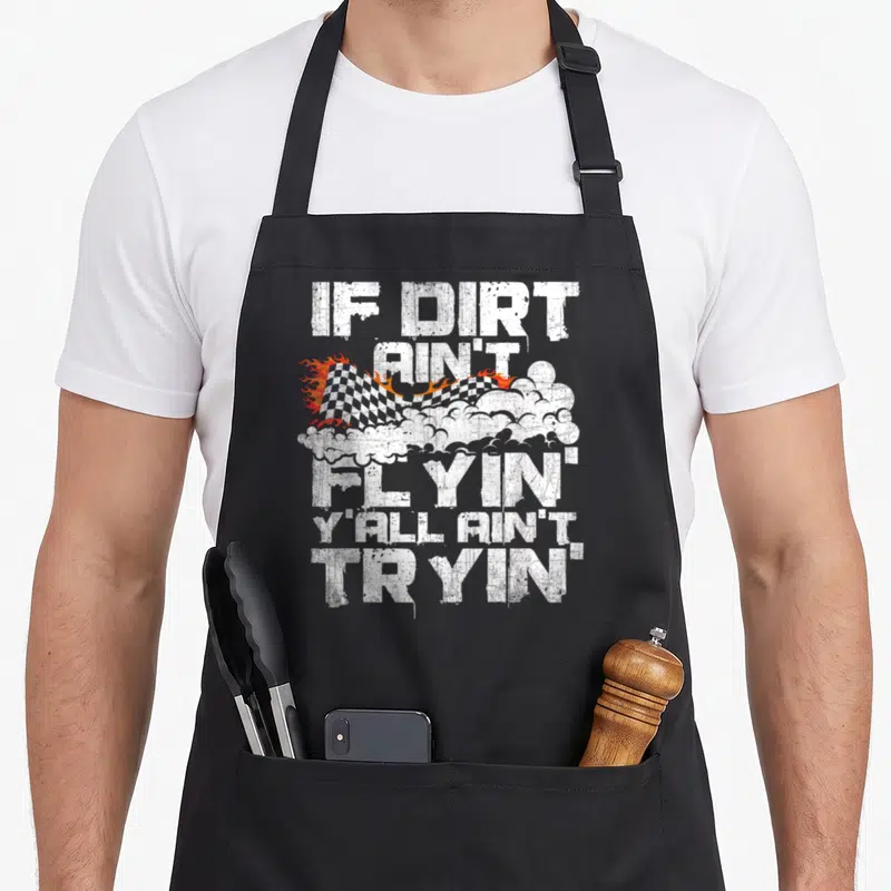 Flyin track apron - Distressed white text IF DIRT AIN'T FLYIN YALL AIN'T TRYIN over checkered flag with fire and dust — shown on a black cotton canvas apron against a white background