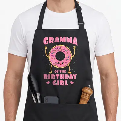 Gramma donut apron - Pink cartoon donut with raised arms and GRAMMA OF THE BIRTHDAY GIRL text flanked by two hearts — shown on a black cotton canvas apron against a white background