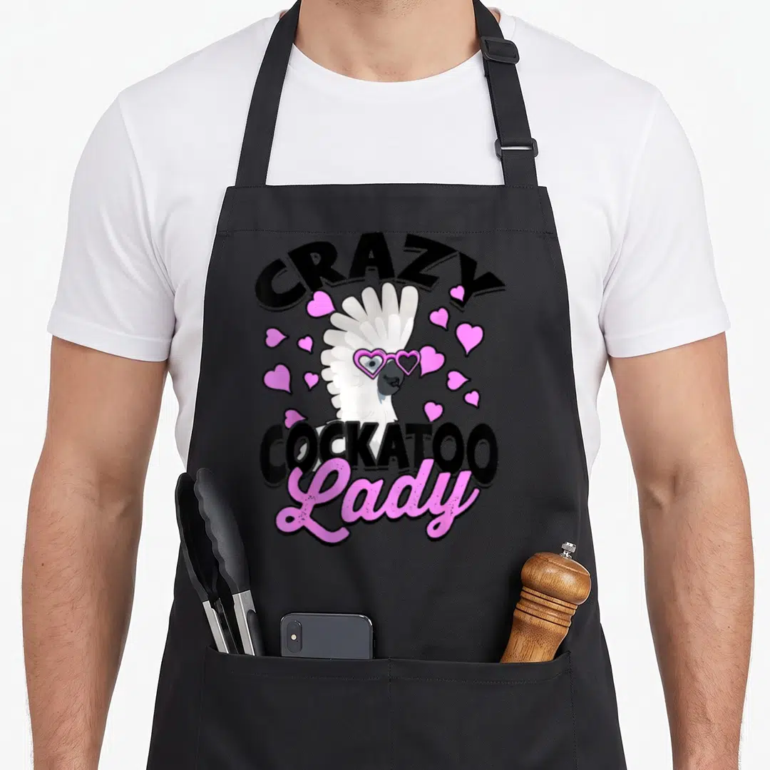 Parrot pet bird apron - Crazy Cockatoo Lady text with white bird wearing pink heart glasses and scattered hearts — shown on a black cotton canvas apron against a white background