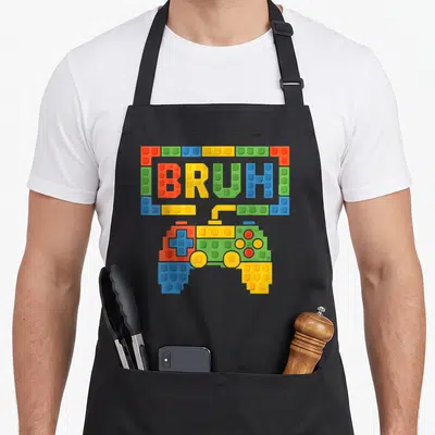 Bruh building aprons - BRUH text in plastic bricks above a brick-built game controller in red, yellow, green, blue — shown on a black cotton canvas apron against a white background