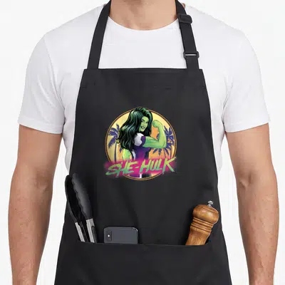 She attorney apron - Cartoon She-Hulk flexing inside yellow circle with palm trees and pink text — shown on a black cotton canvas apron against a white background