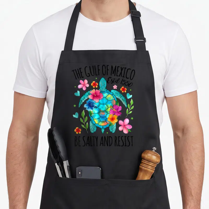 Estd mexico apron - Sea turtle with pink flowers hearts and text THE GULF OF MEXICO Estd 1550 BE SALTY AND RESIST — shown on a black cotton canvas apron against a white background