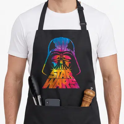 Helmet tie apron - Tie-dye Darth Vader helmet silhouette with STAR WARS text in blue red yellow and purple — shown on a black cotton canvas apron against a white background