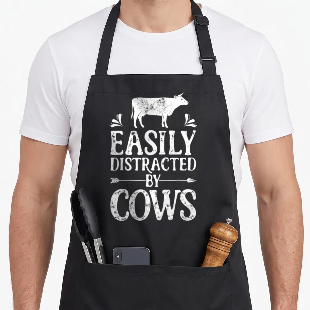 Cow farmers apron - Distressed white cow silhouette above stacked text EASILY DISTRACTED BY COWS with horizontal arrow — shown on a black cotton canvas apron against a white background