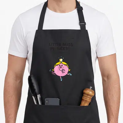 Miss princess apron - Pink round character with yellow hair and crown holding a wand under stacked text — shown on a black cotton canvas apron against a white background