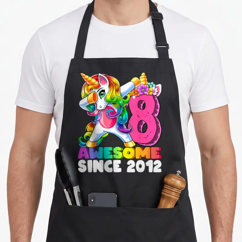Unicorn 8th birthday apron - Cartoon unicorn dabbing next to pink number 8 with rainbow text — shown on a black cotton canvas apron against a white background