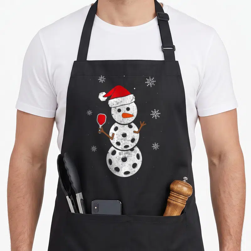 Santa snowman apron - White snowman with hole-patterned body holding red paddle and wearing red hat among six snowflakes — shown on a black cotton canvas apron against a white background