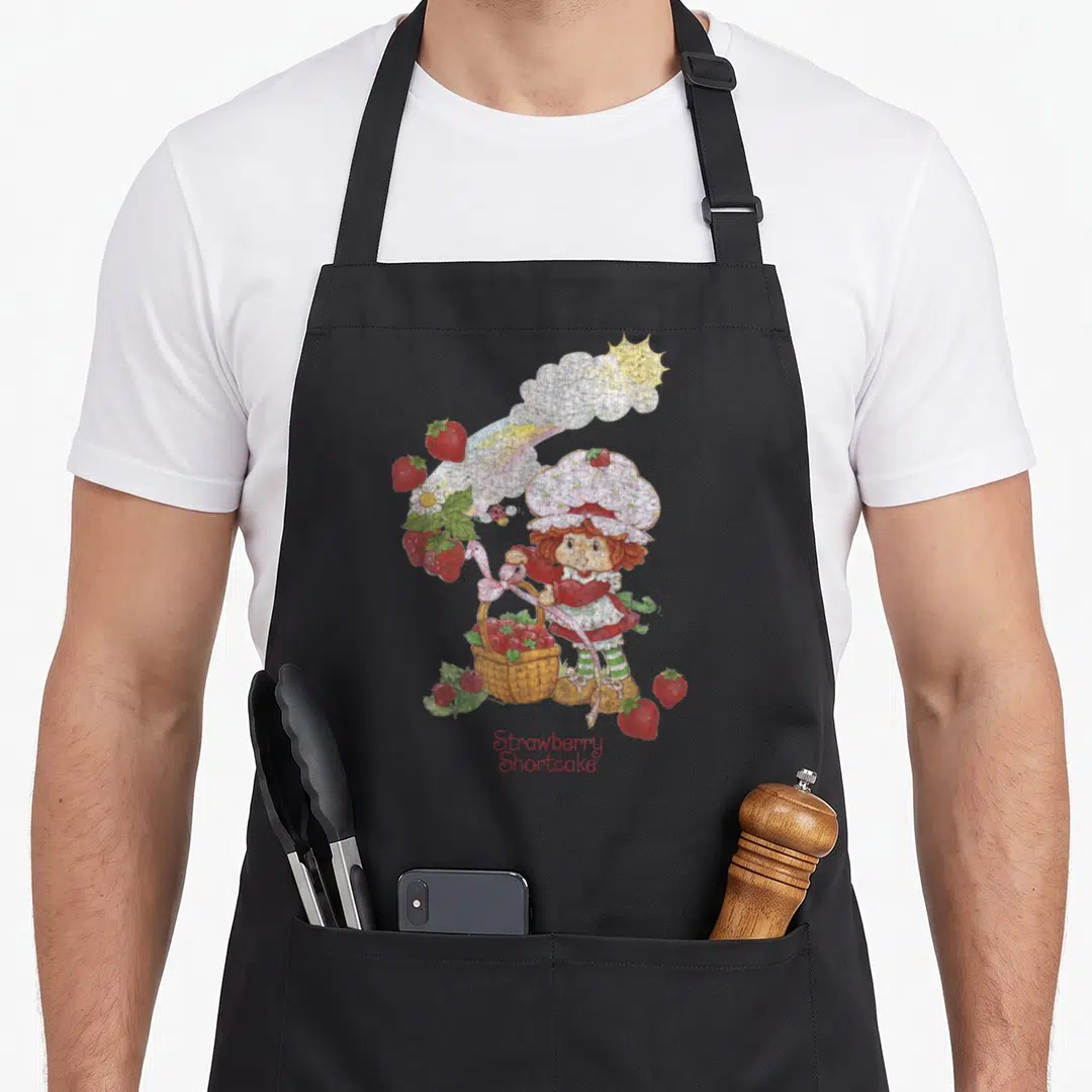 Rainbow strawberry apron - Cartoon girl holding red strawberry basket under rainbow and sun with printed text below — shown on a black cotton canvas apron against a white background