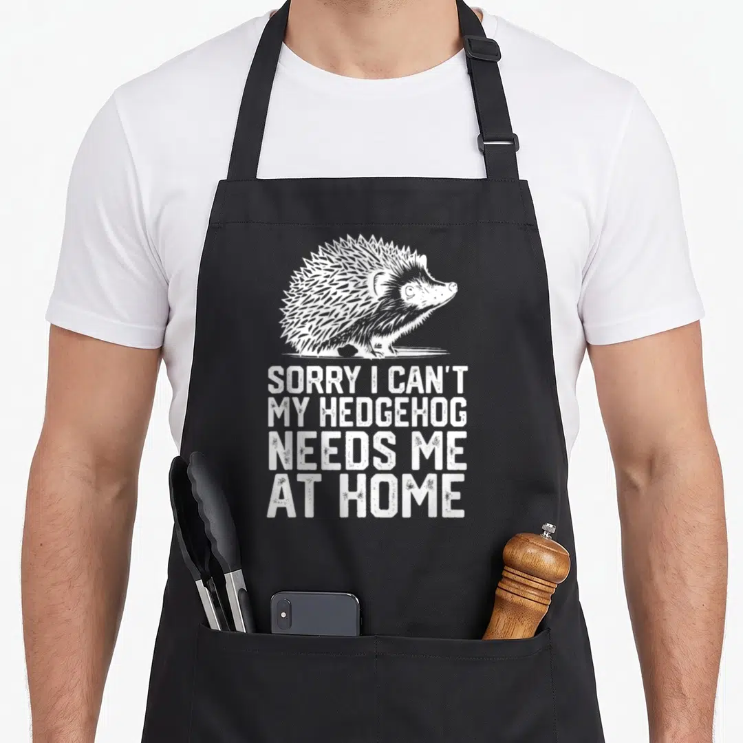 Hedgehog at home aprons - White distressed hedgehog illustration above four stacked lines of text on black fabric — shown on a black cotton canvas apron against a white background