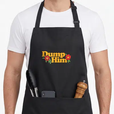 Dump sage apron - Yellow Dump Him text with two red flowers and green leaves in cartoon style — shown on a black cotton canvas apron against a white background