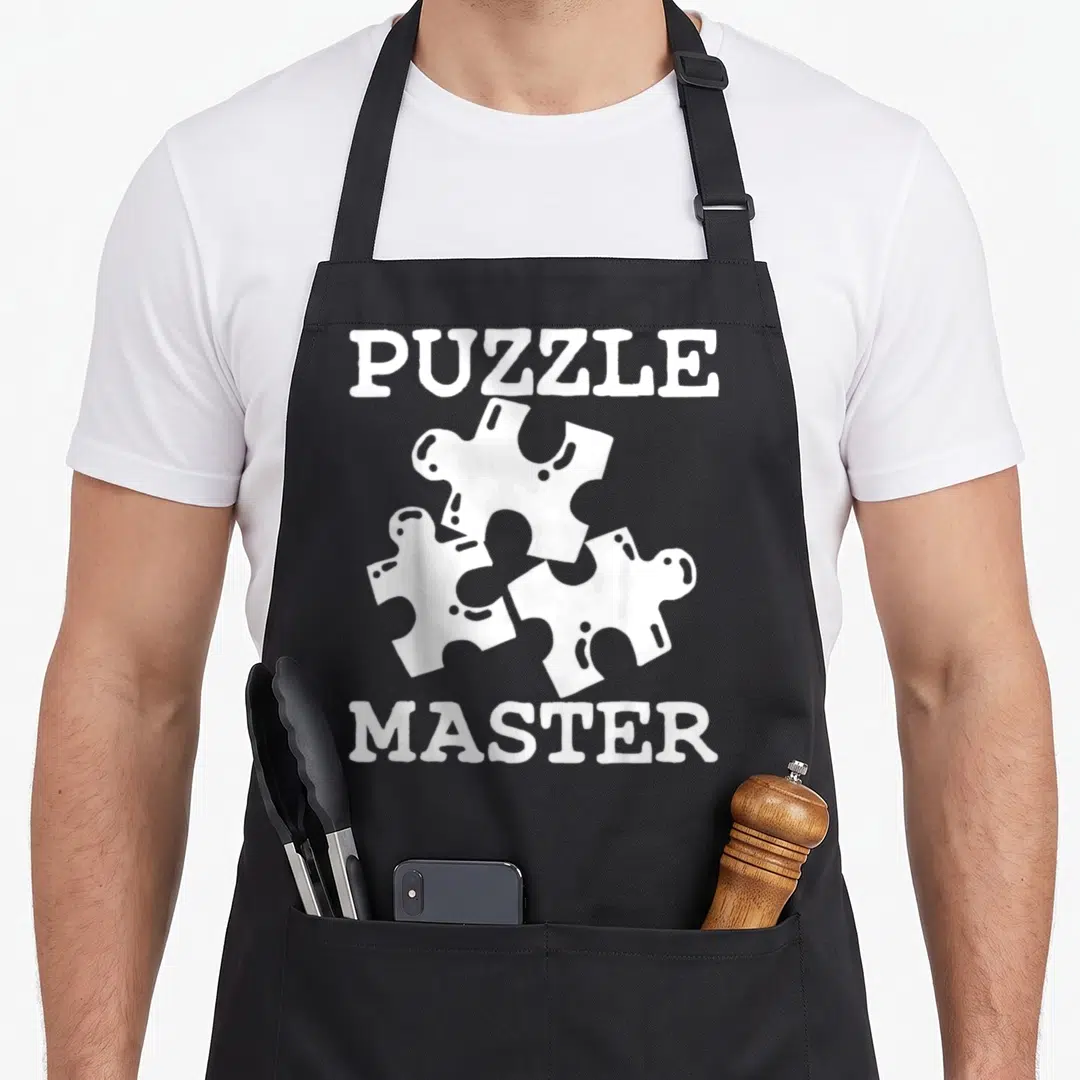 Puzzle master apron - White PUZZLE MASTER text surrounding three white jigsaw puzzle pieces — shown on a black cotton canvas apron against a white background