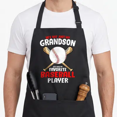 Hes baseball apron - Crossed tan bats behind white baseball with red stitching and stacked text in red and white — shown on a black cotton canvas apron against a white background